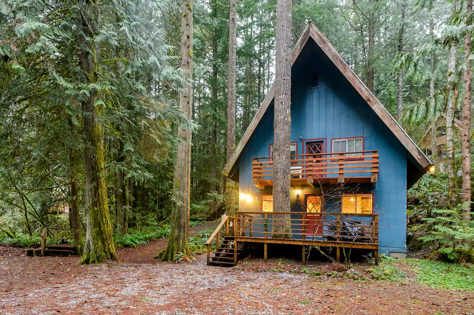 Photo showing the outside of this two story cabin, which is painted blue. There is an upstairs and a downstairs deck. The cabin is surrounded by evergreen trees and one other cabin is just faintly visible behind it. 