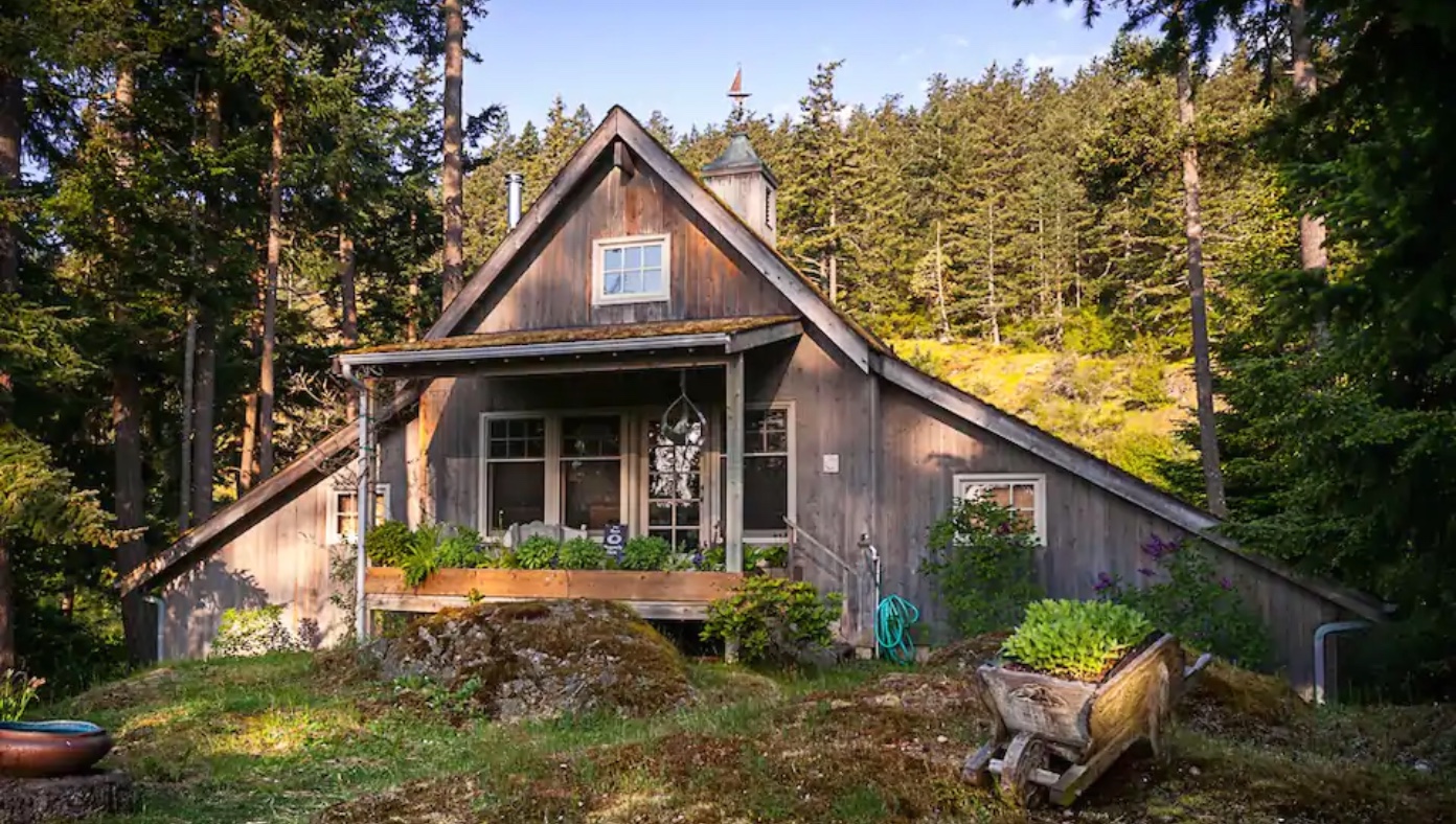 Image of the exterior of the refurbished barn. Plants grow in a wooden wheelbarrow out front. The cabin is surrounded by hills and evergreen trees. This is one of the best cabins in Washington State.
