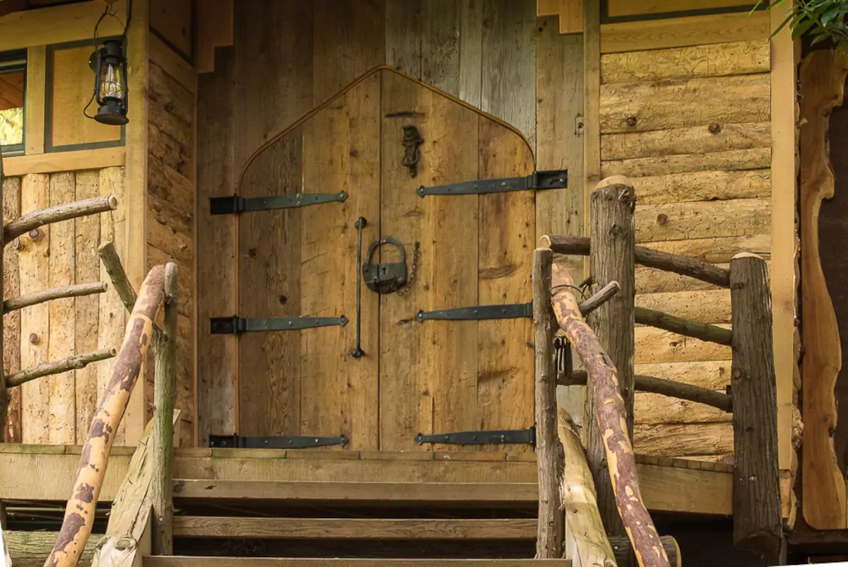 Photo of wooden stairs leading up to a log cabin with castle-like front doors. Front doors have iron hinges and hardware. A lantern hangs beside the front door. This is one of the most unique cabins in Washington State. 