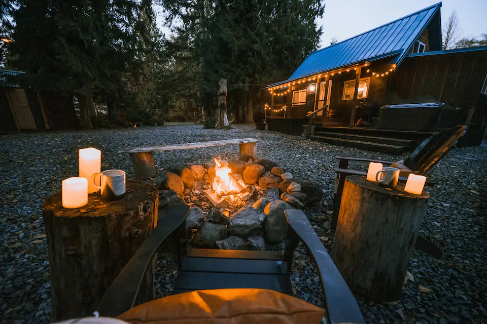 Photo of a fire pit surrounded by cozy chairs and candles. The cabin is in the background with inviting strings of lights on the front porch. Cabin 135 is one of the best cabins in Washington State. 