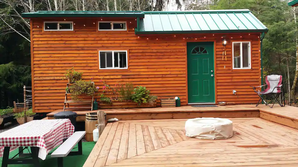 Towable trailer sized tiny house with cedar siding, a green roof and matching door. Large patio, picnic table, and fire pit are visible in this photo. The Rialto tiny house is one of the best cabins in Washington State. 