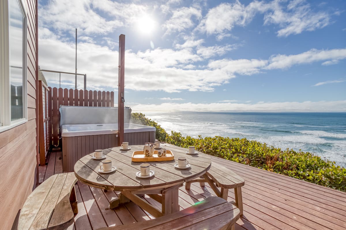 The private deck of a beach house in Oregon with a large hot tub, a patio table, and endless views of the Pacific Ocean