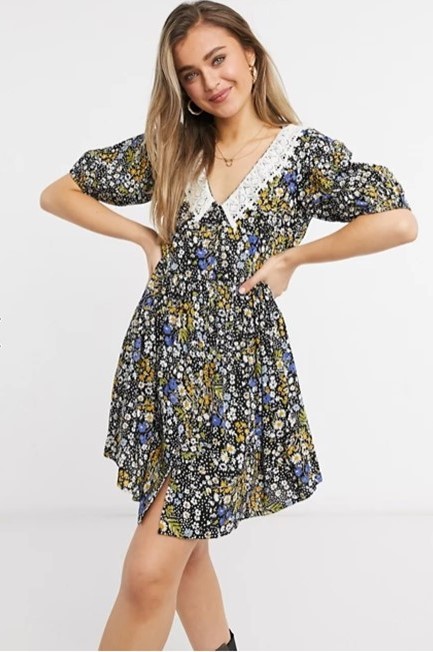 A woman wearing a mini smock dress that is black floral with white, blue, and yellow flowers. It also has v-neck with a crochet peter pan collar.