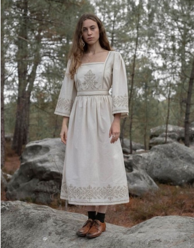 A woman dressed in a white cotton dress with cream embroidery, long sleeves, and midi length hem, standing on a rock in the woods