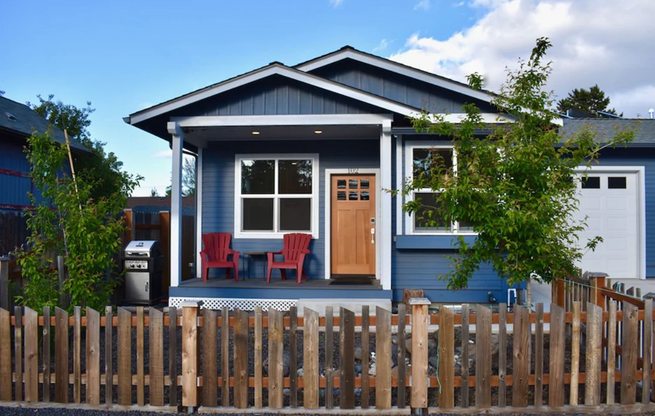 The exterior of a charming cottage in Bend that is painted dark blue, has a small private porch, and a natural wood front door. 