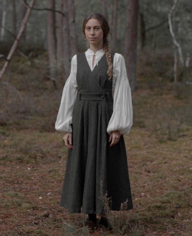 A woman standing in the woods wearing a charcoal grey wool dress and a cream colored embroidered tunic under it.