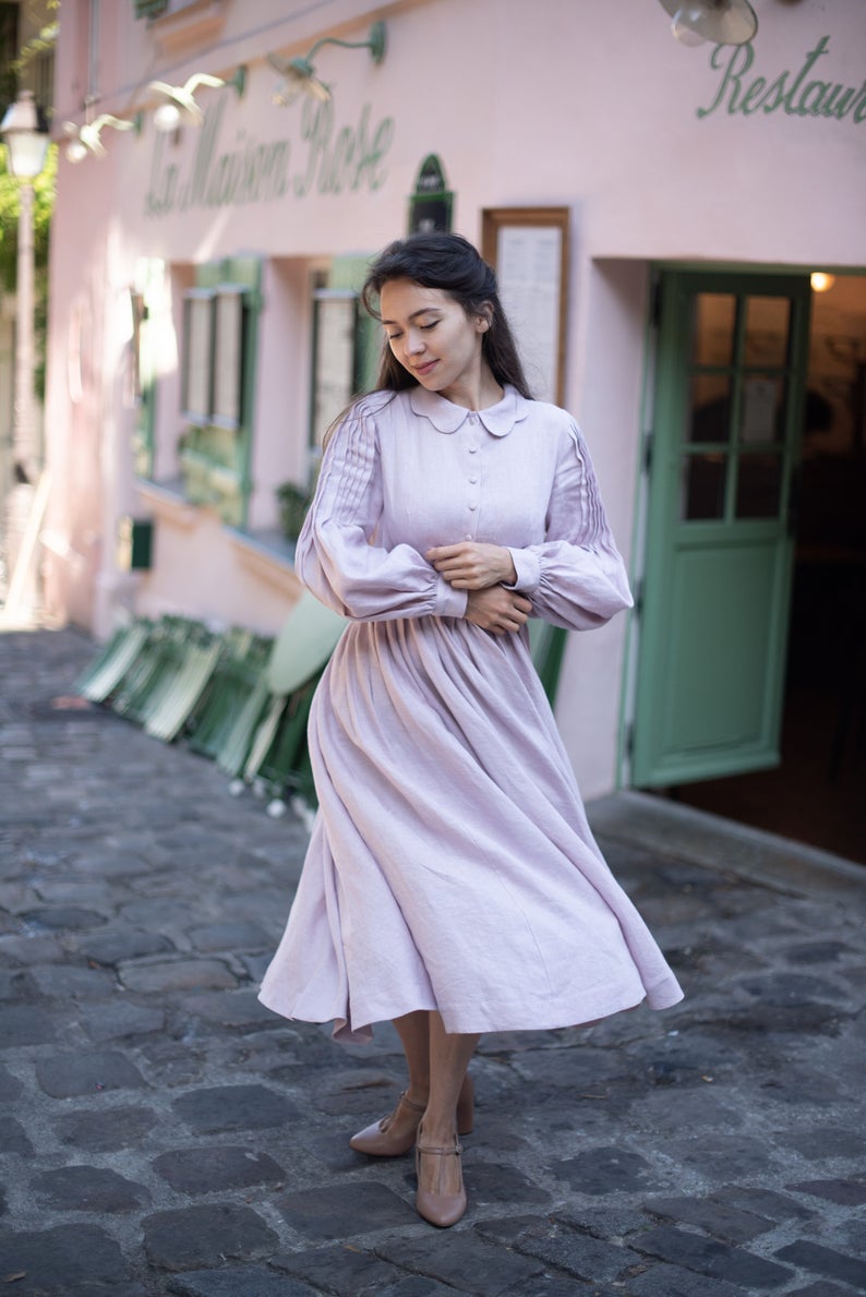 A woman in a dusty rose pink cottagecore dress with long sleeves, a scalloped peter pan collar, and buttons up the front of the chest on a cobblestone street