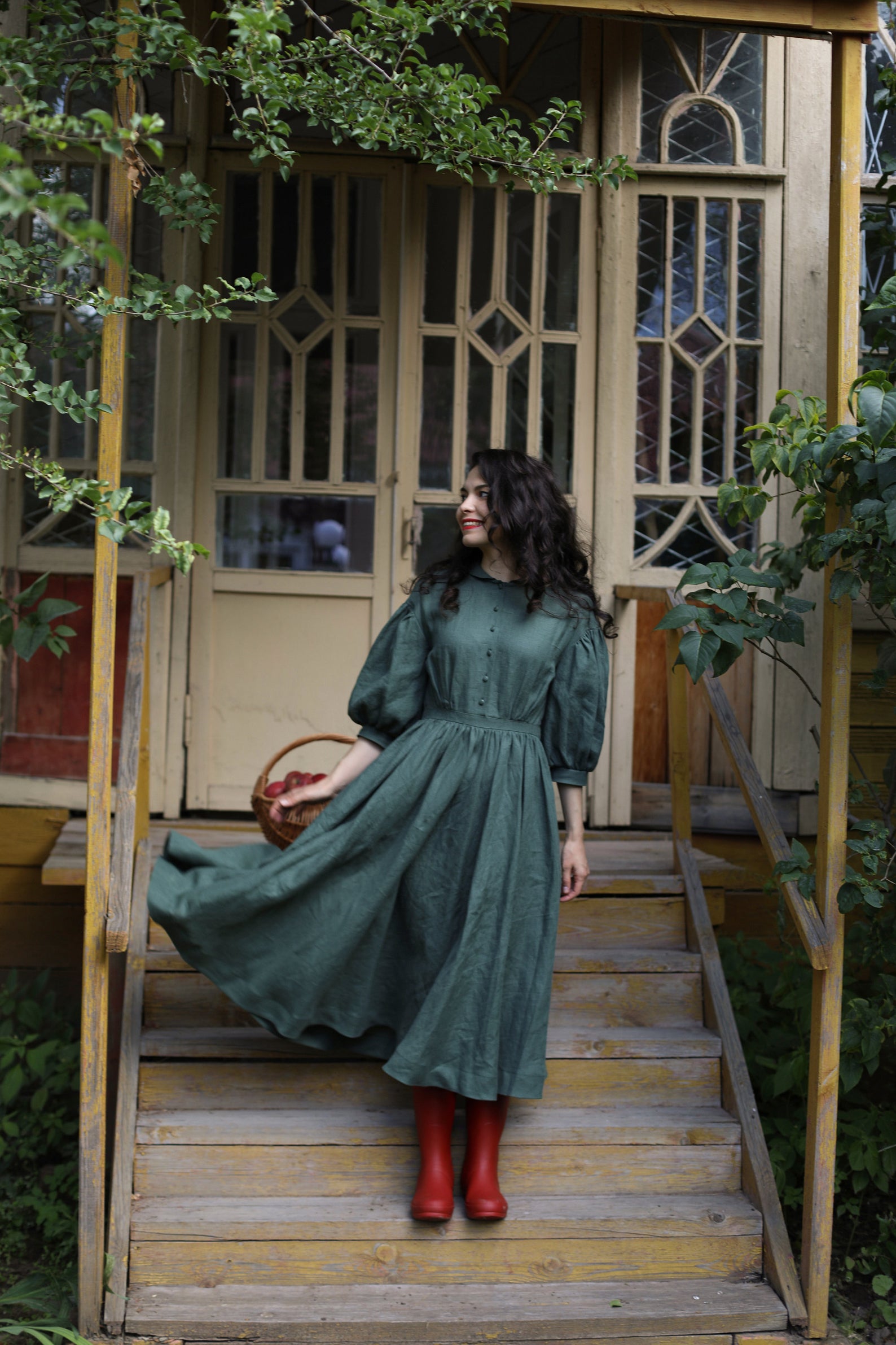 A woman standing on steps in a dark green linen dress with puffy sleeves, buttons down the front, and dark red rainboots. 