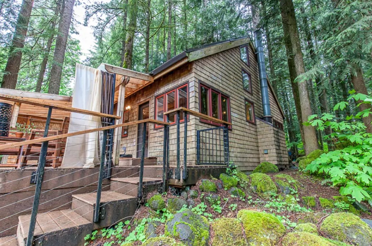 The exterior of a wood shingle cabin in the middle of the woods surrounded by lush greenery with a large deck