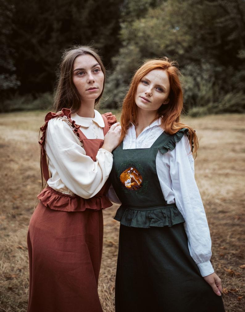 Two women wearing the same apron dress in two different colors that has a sleeping fox embroidered on the front of it.