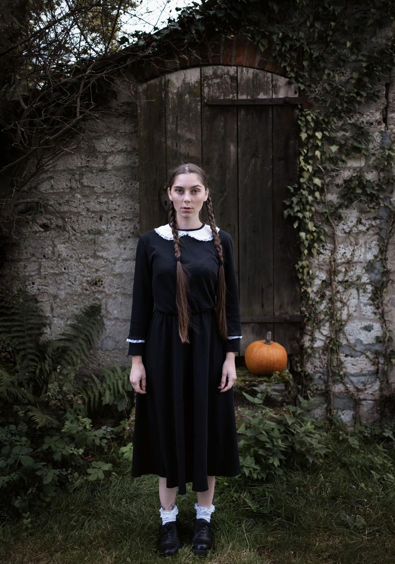 A woman standing in a stone courtyard with greenery around her wearing a black linen dress with a white peter pan collar and lace on the wrists of the long sleeves with a pumpkin behind her best cottagecore dresses