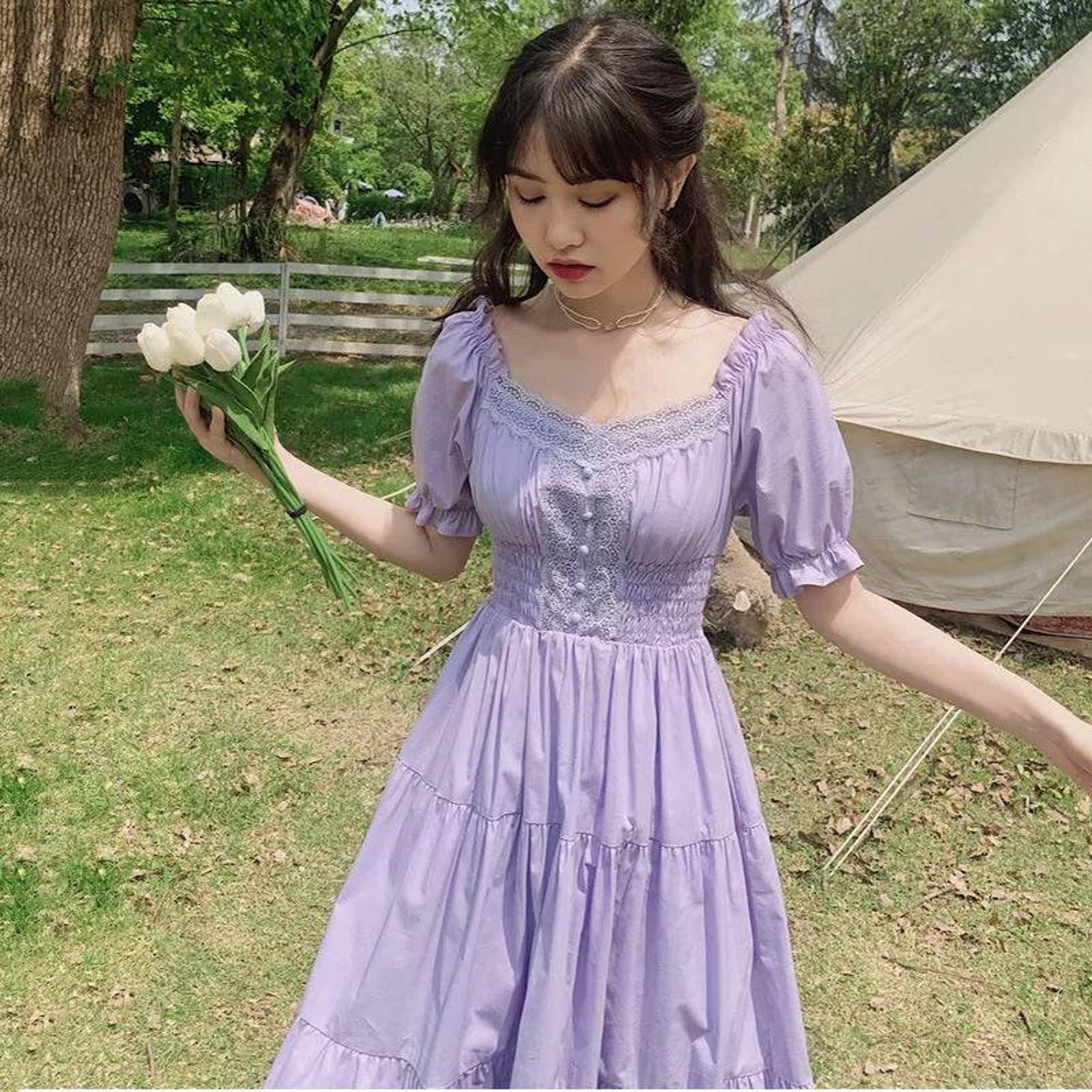 A woman on a farm holding flowers and wearing a pastel purple dress with puffy sleeves, lace, and a tiered skirt one of the best cottagecore dresses