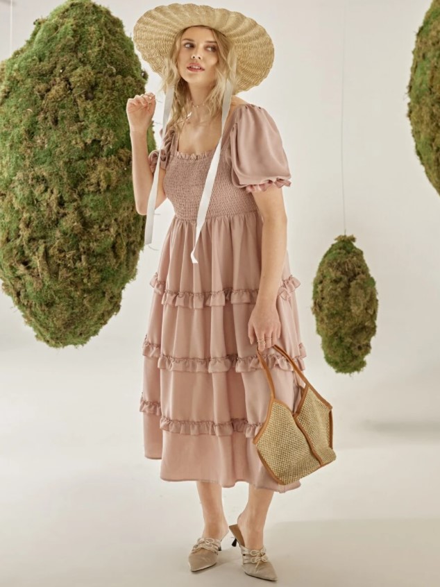 A woman wearing a dusty pink ruffled dress, a sun hat, and a boho purse in a space with topiaries around her