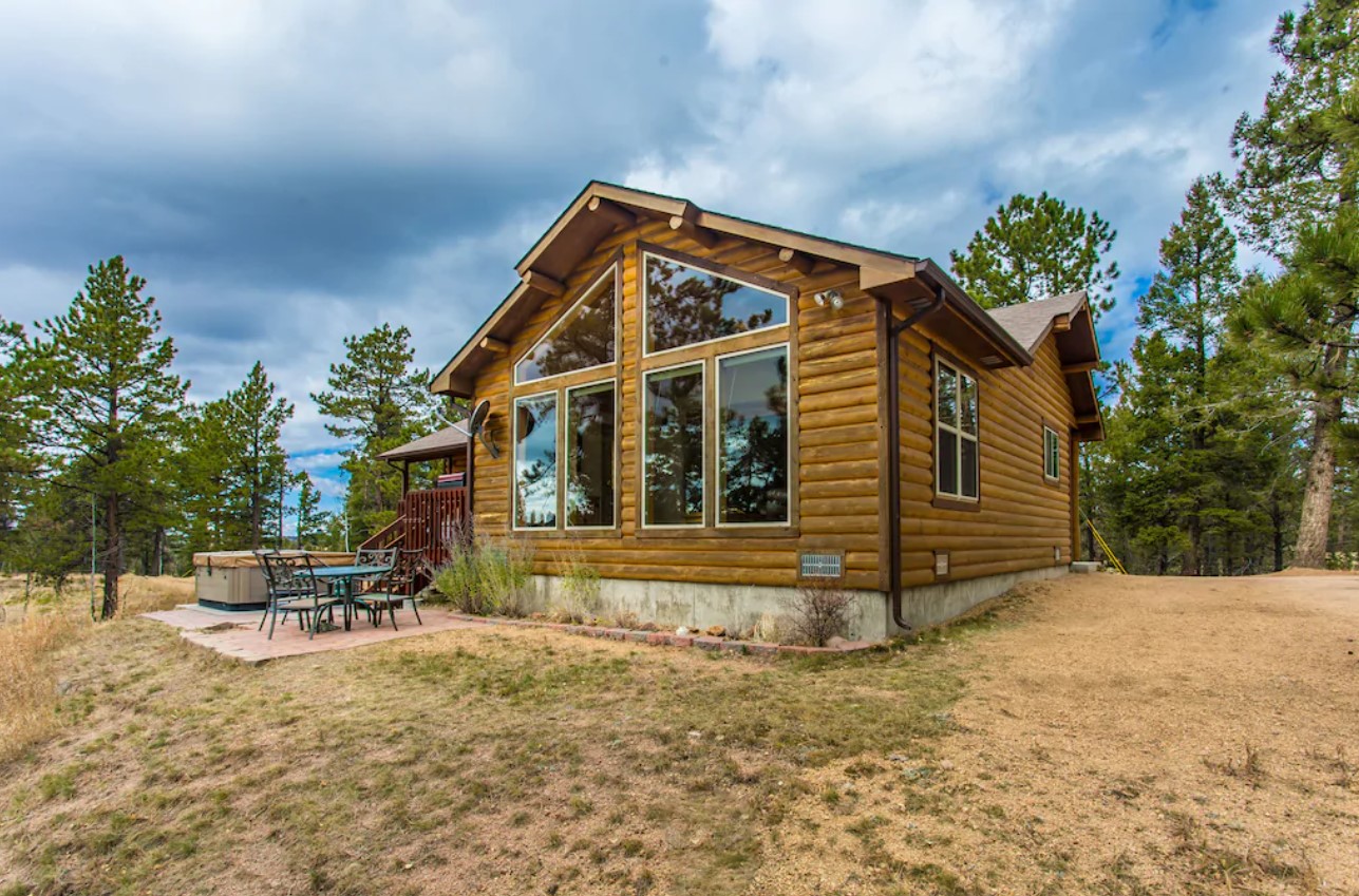 A modern log cabin with plenty of windows, a nice patio, and a hot tub, on a cloudy day