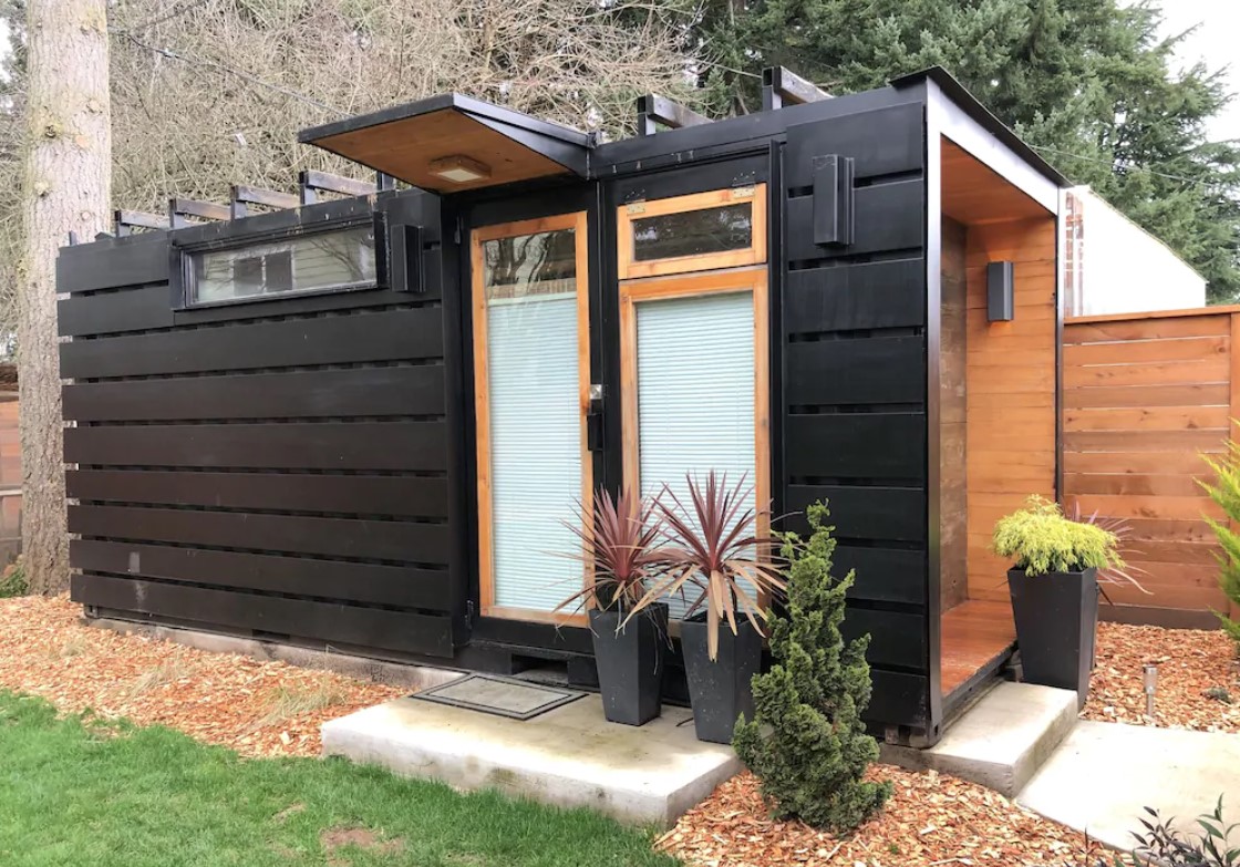 The exterior of a shipping container turned modern tiny home that is painted black and has plants around it