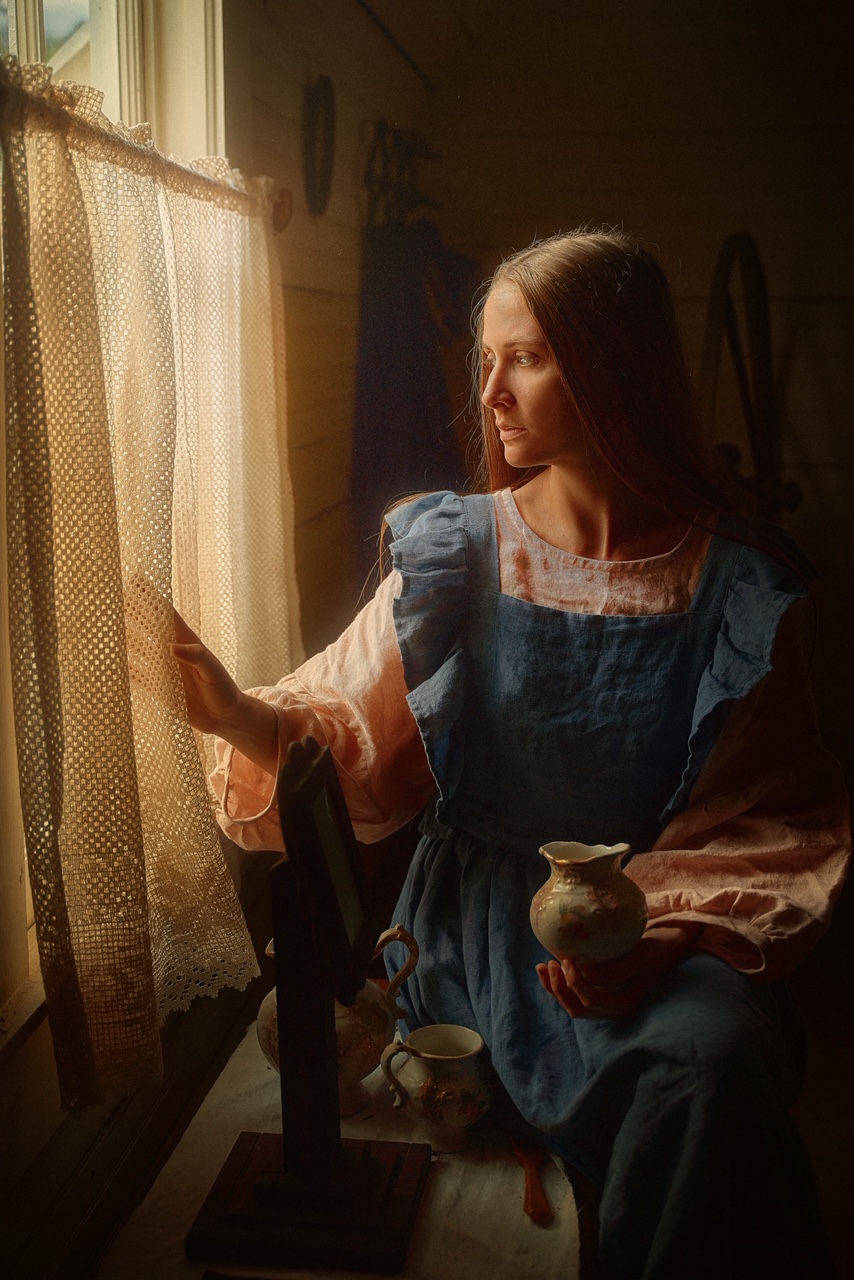 A woman sitting by a window wearing a pink tunic and blue sleeve ruffle dress