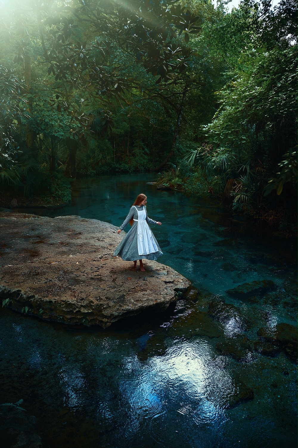 A woman standing by water wearing a grey linen dress and white apron