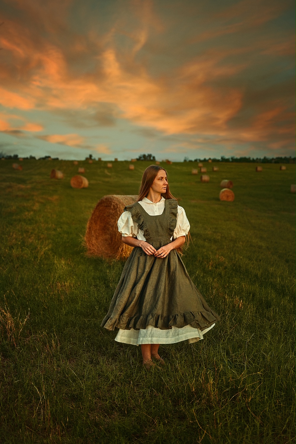 A woman standing in a field of stacked hay wearing a cottagecore dress and pinafore