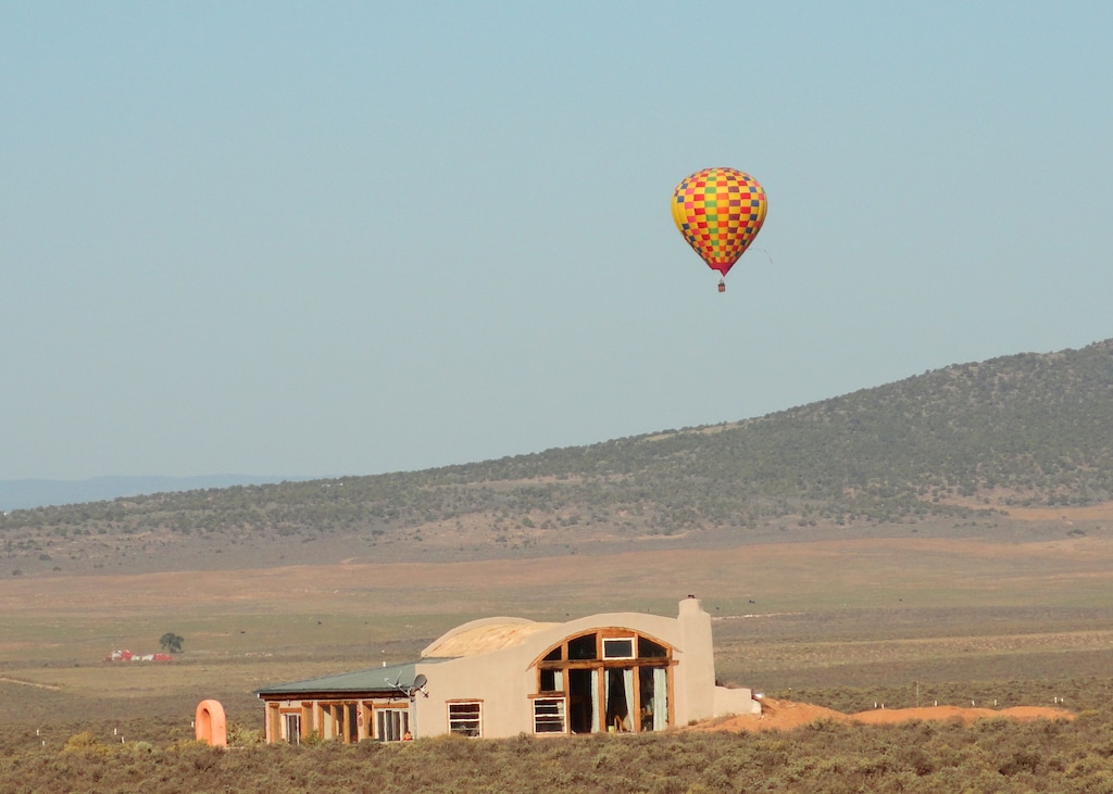 A clay home with large windows and a domed roof line in the desert of New Mexico. There is nothing around it except for a slight hill in the background. Floating near the home in the sky is a hot air balloon with red, green, orange, blue, and purple checked fabric.