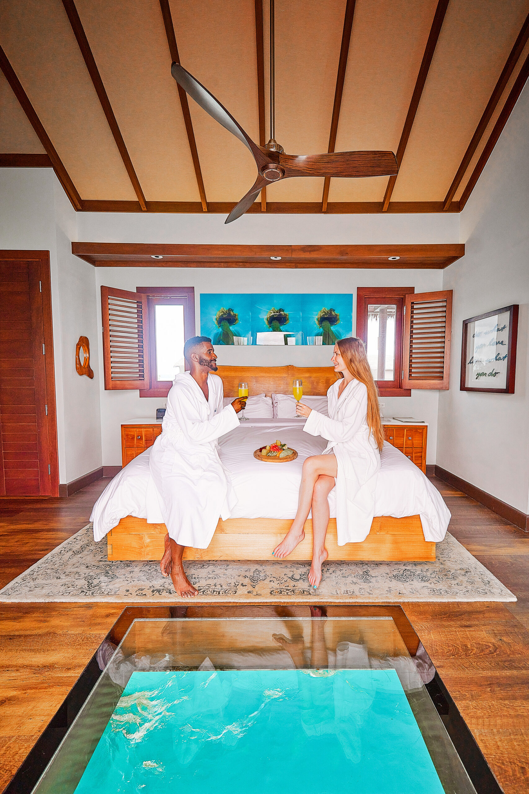 A couple sitting on a bed enjoying mimosas and fruit in their robes with an window to the ocean near their feet