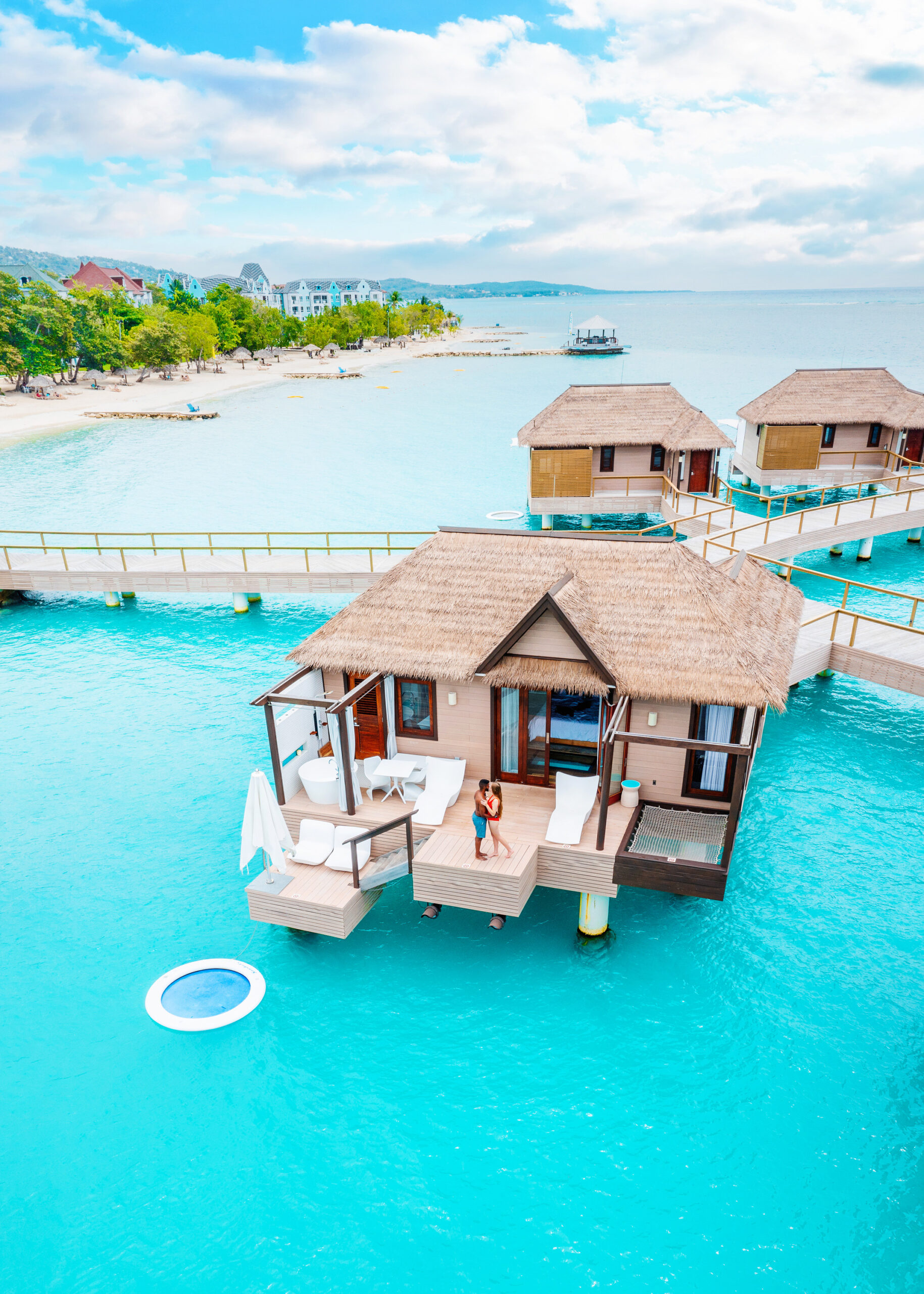 A couple standing on the deck of an overwater bungalows in Jamaica where there are sun loungers, a tub, a bistro set, an outdoor hammock, and a floating ring