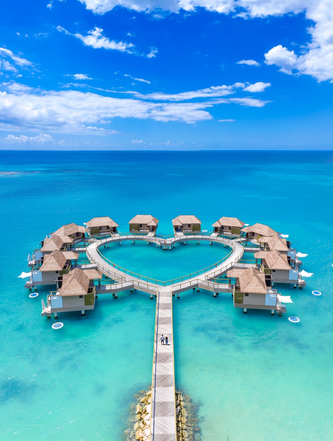 an aerial photo of two people standing on the dock facing a heart shaped collection of overwater bungalows in Jamaica