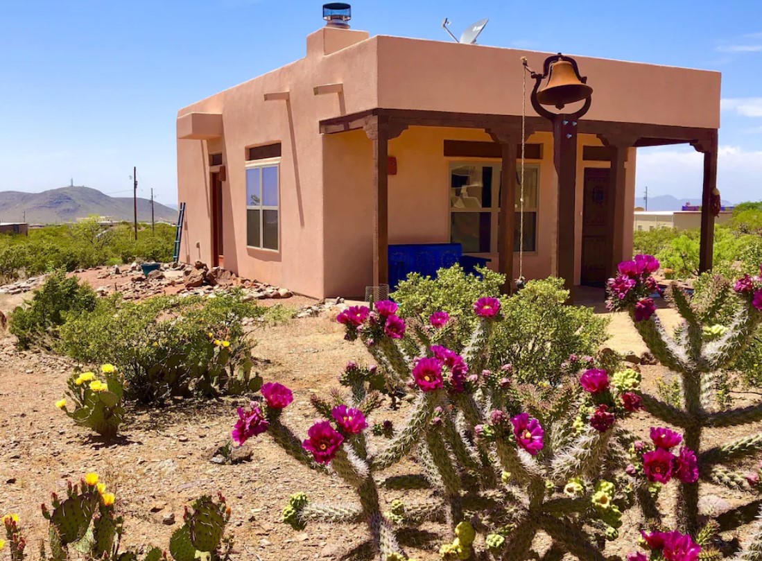 A house made out of red clay with dark wood accents and a covered front porch. There is a standing bell in front of the house, as well as yellow and pink desert flowers