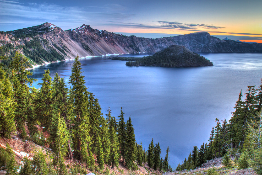 View of the caldera of Crater Lake at sunrise. Wizard Island is visible and the sunset is bringing out the purple tones of the sloping shores of the lake. 