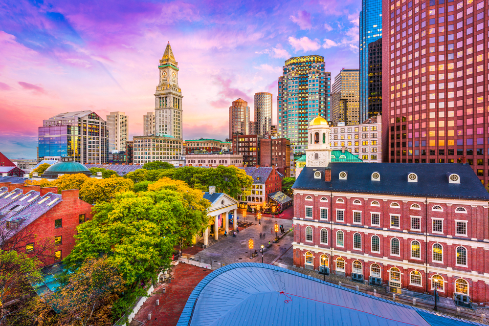 The skyline of Boston, Massachusetts at sunset. You can see very old buildings mixed with modern skyscrapers. The sky is blue, pink, and purple.