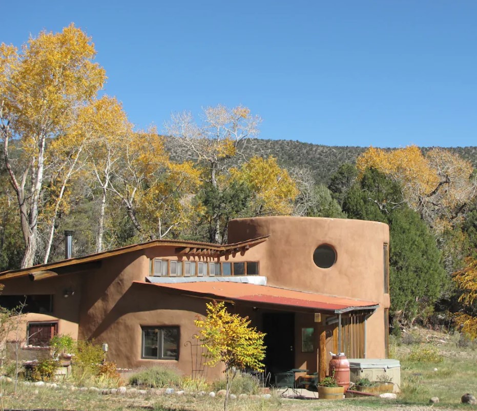 A uniquely shaped adobe clay home in a mountain valley with a mountain in the distance and trees with green and yellow foliage on them.