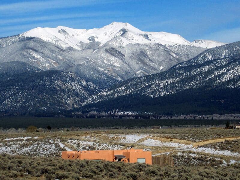 A large red clay home in a valley in the mountains in New Mexico. The mountains are snow peaked, but there is no snow near the home. It has a desert landscape with short shrubs and sandy areas.