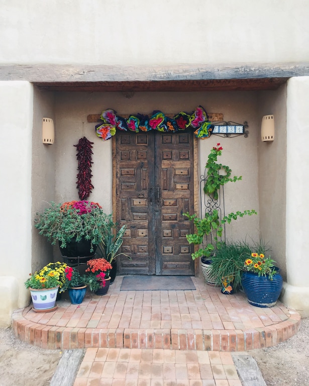 The street entrance to Casa Azul, a unique home in Mesilla Plaza New Mexico. It has two large wooden doors, and several potted plants. The plants have yellow, pink, and orange flowers. Over the door is an arrangement of paper flowers. Airbnbs in New Mexico