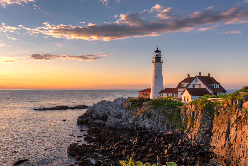 A lighthouse on the coast of maine's rocky shore at sunset. It is a large white lighthouse with large white buildings near it.