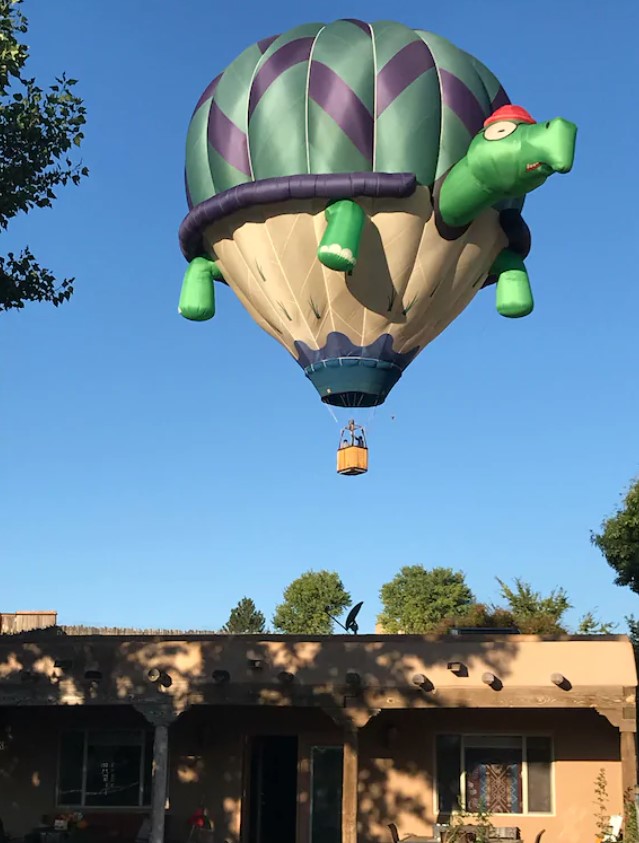 A hot air balloon that looks like a turtle wearing a red hat and glass flying over a clay adobe home. The hot air balloon has arms, a green shell with purple stripes, and a tan 'underbelly'. It is floating very close to the adobe style home