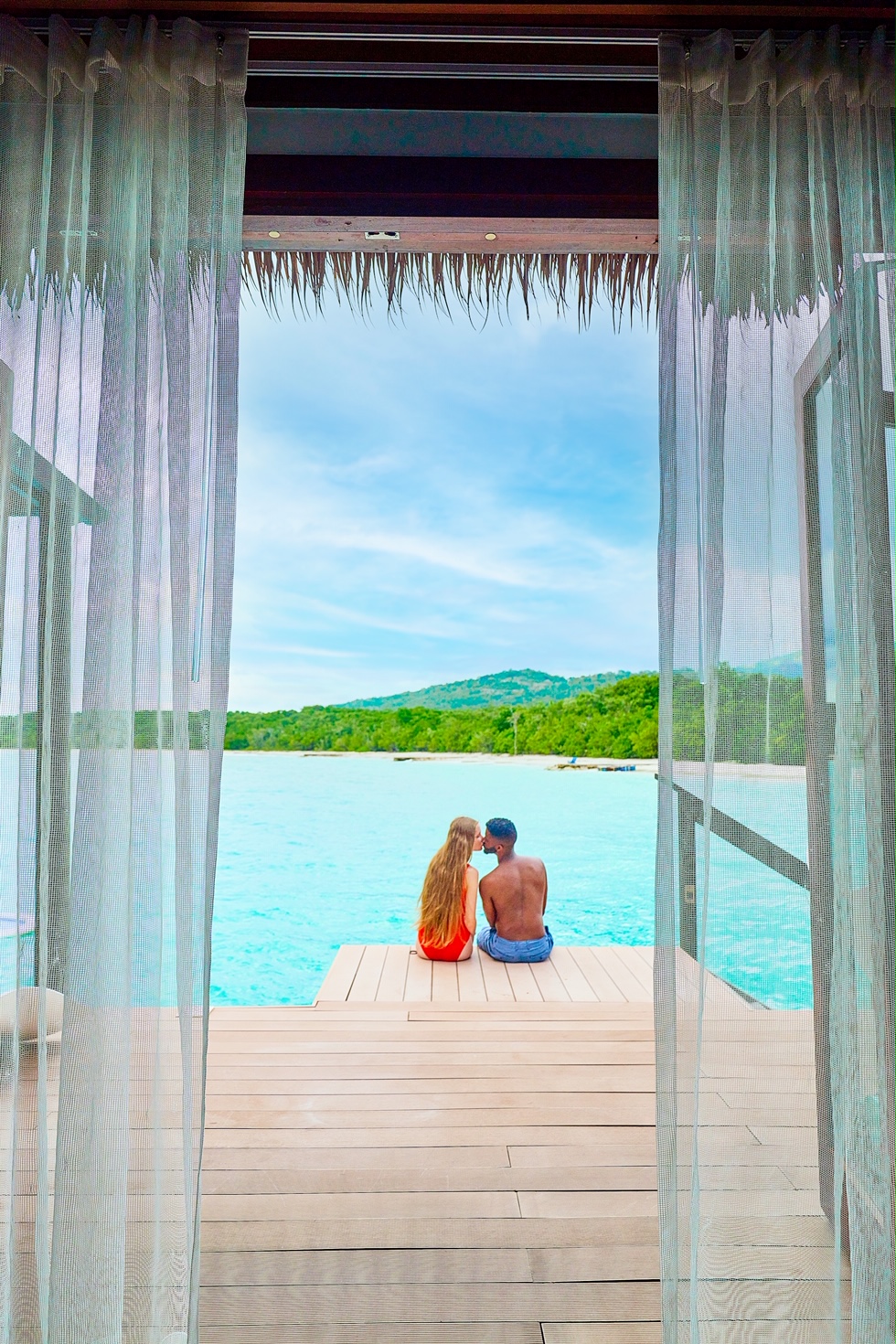 A couple sitting and kissing on the deck of an overwater bungalow with a view of the ocean and mountains