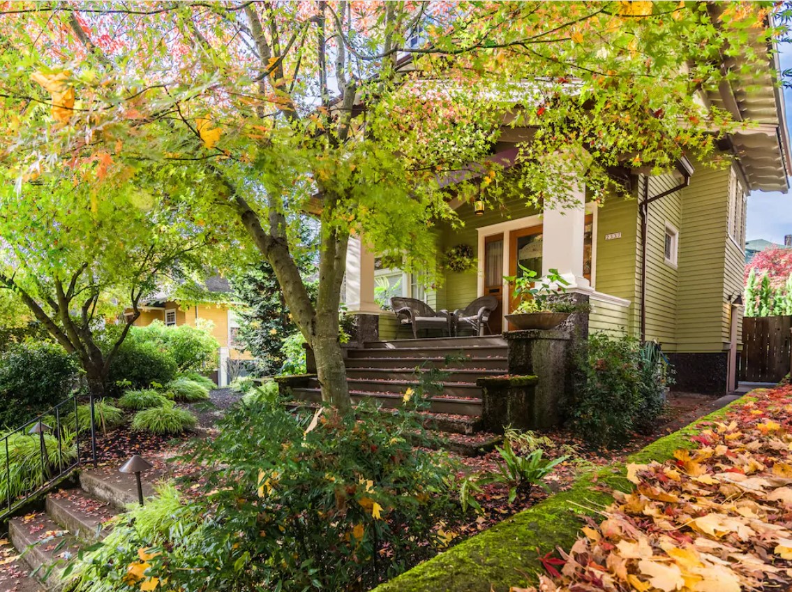 The exterior of a pea green Craftsman-style home with a large front porch and stone steps. There are large trees and shrubs in front of the house. It is one of the best vacation rentals in Oregon.