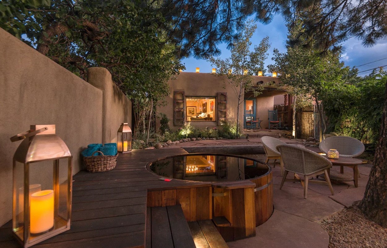 The backyard of a historic adobe in Santa Fe New Mexico. It has a large stone patio with seating, a cedar plank hot tub, and lots of lights. The home is in the back with a big window and a small deck with two rocking chairs. Airbnbs in New Mexico