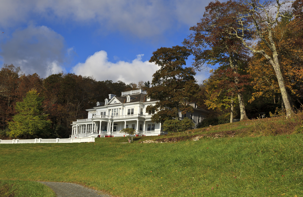The Moses H. Cone Manor on the Blue Ridge Parkway surrounded by a grassy lawn and large old trees. The sky is very blue with big fluffy clouds. Its a great stop on any east coast road trip.