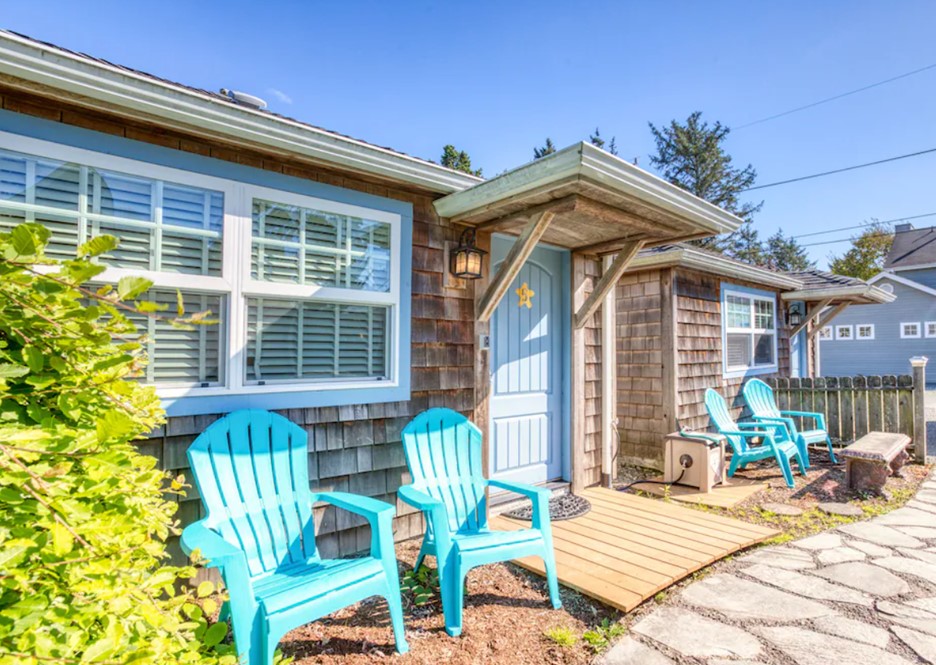 The exterior of a raw wood shingled cottage with a pale blue door, pale blue trim, and bright blue Adirondack chairs in front of it with a bush in the corner on a sunny day.