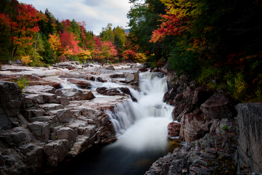 the Rocky Scenic Gorge in New Hampshire which is a waterfall. next to the river and gorge are trees with leaves changing colors. The leaves are yellow, red, and orange.