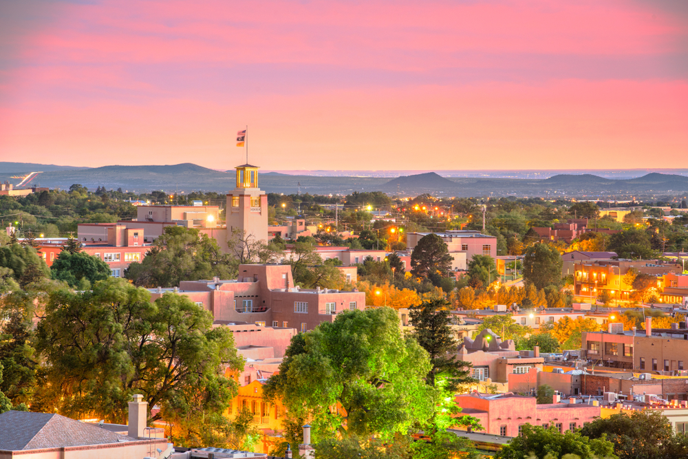 A view of Santa Fe New Mexico at sunset with a pink sky, the plaster buildings tinted pink from the sky, and the buildings lit up