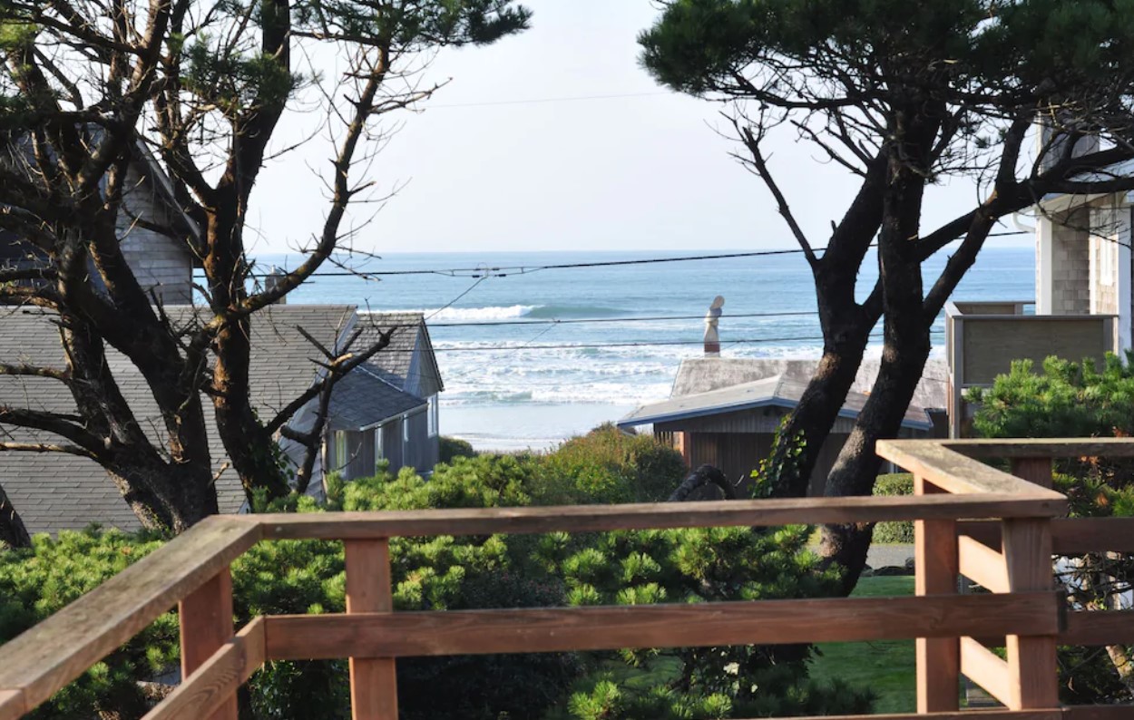 The view of the ocean from the deck of an Oregon vacation rental with houses in the distance, and trees and shrubs near the deck