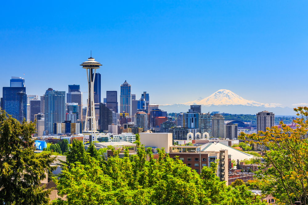 The Seattle skyline with the space needle in the front and a mountain in the background on a sunny day.