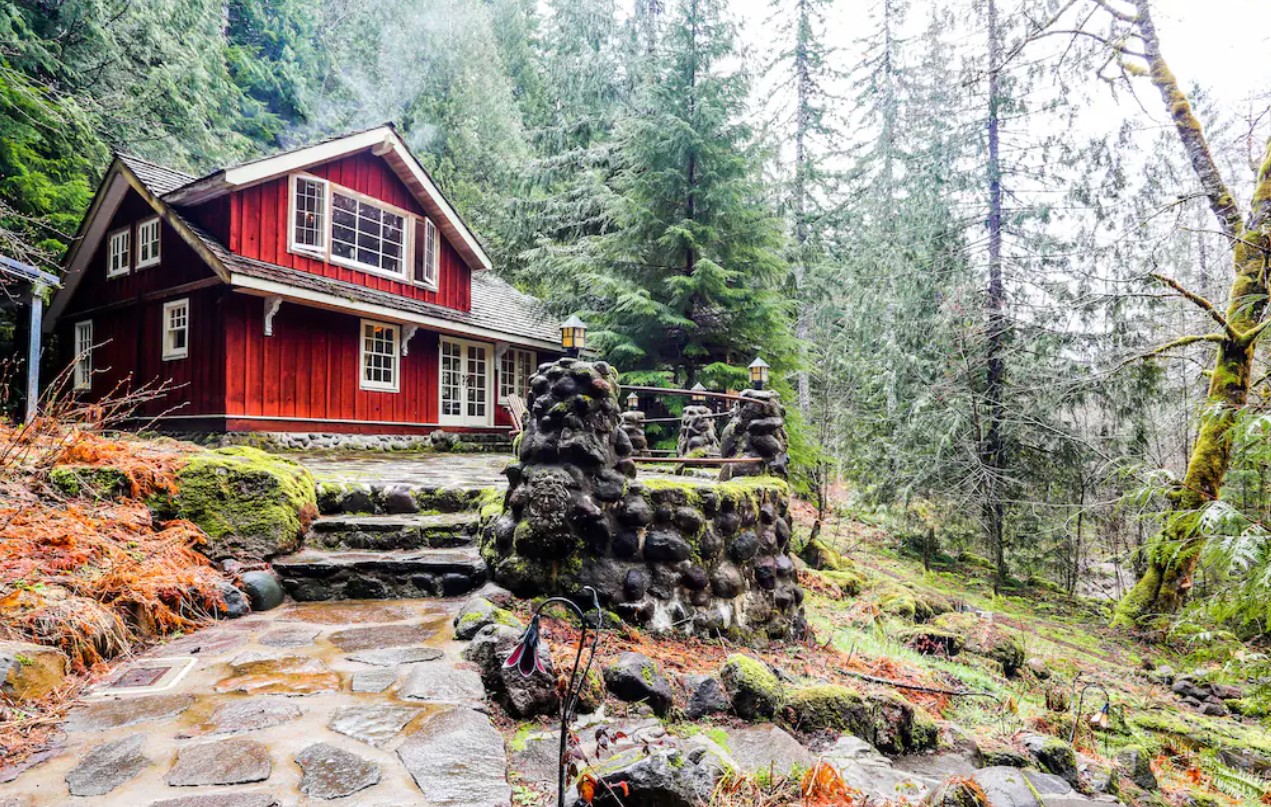 The exterior of a bright red painted lodge in the woods surrounded by trees with stone steps leading to a stone patio in front of the lodge house one of the best vacation rentals in Oregon