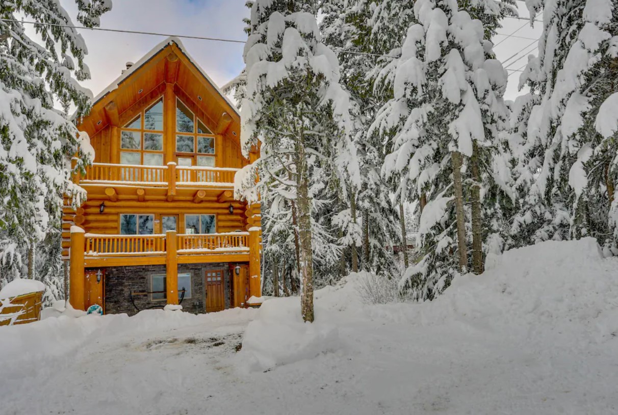 The exterior of a two story wood lodge with two decks and plenty of windows, surrounded by snow and trees covered in snow