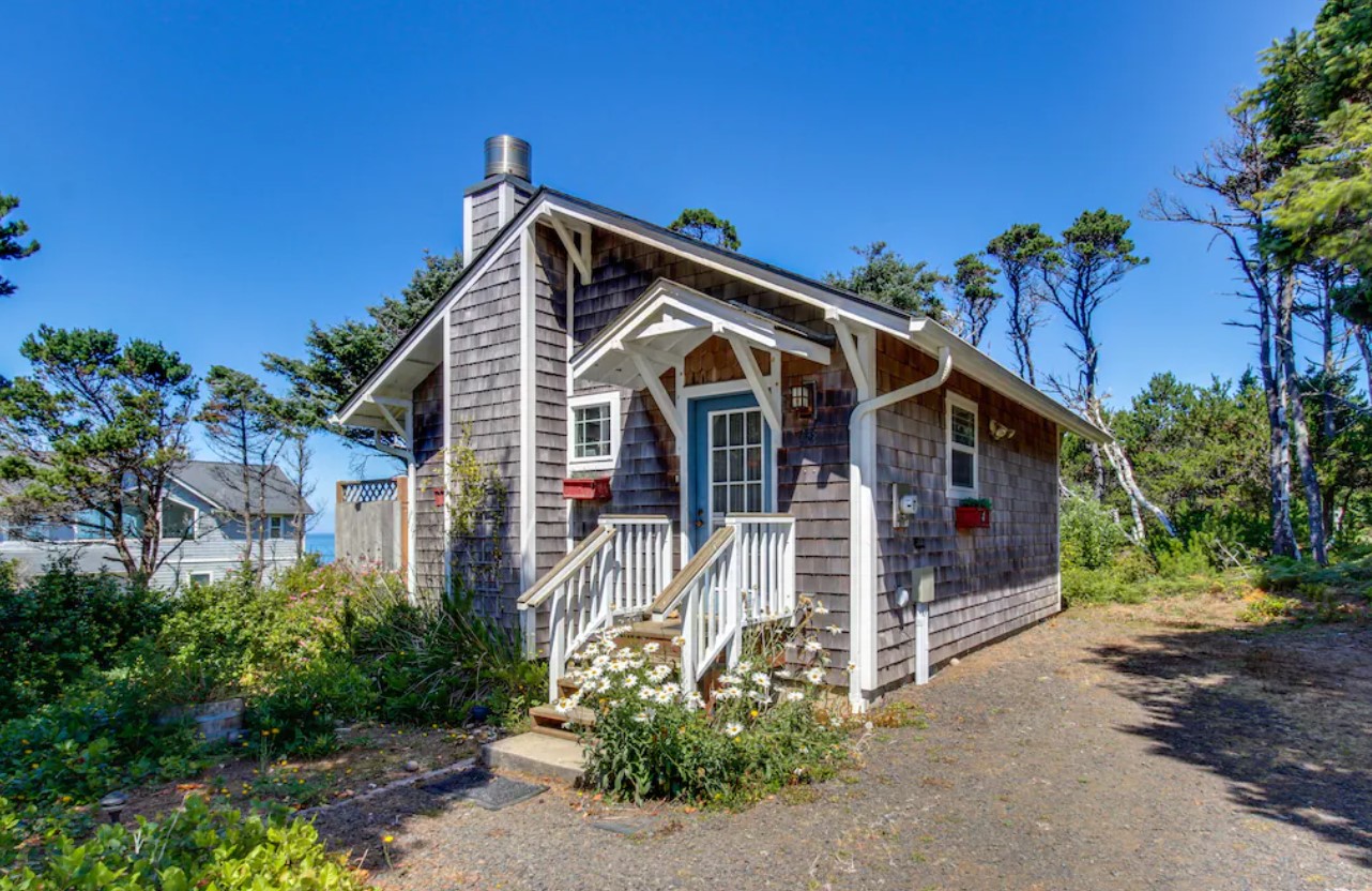 The exterior of a charming beach cottage that has grey shingles, a bright blue door, white trim, and a chimney. In front of the house, there is a small garden with white flowers and shrubs. It is one of the best vacation rentals in Oregon.