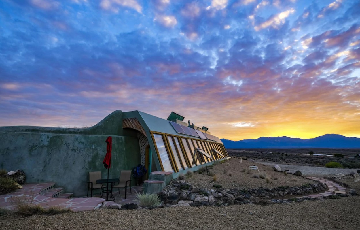 The exterior of an earthship in the New Mexico desert. It is painted a sage green, and has windows all along one side of it. There is also a small patio with a table and chairs and desert mountains in the distance. It is sunset. 