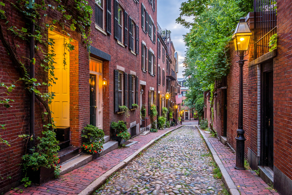 A classic New England Boston cobblestone street. There are antique brick buildings on either side of the cobble stone street, antique style street lamps, and greenery in front of the doors. The windows have black shutters.
