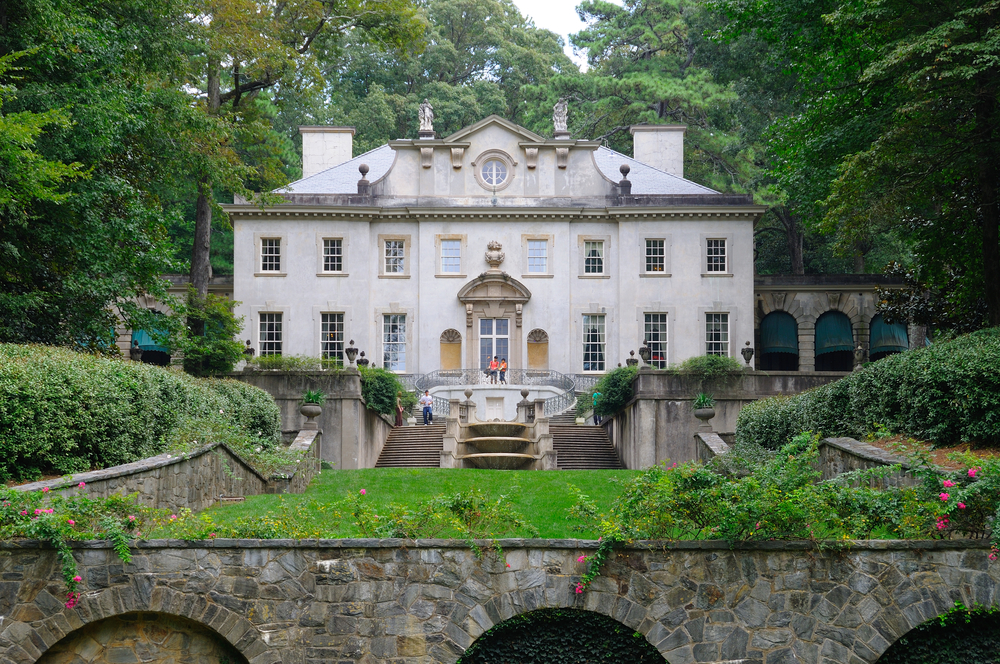 An old manor named the Swan House in the middle of Atlanta. It is a colonial greek revival style home that is cream with lots of windows. There is a lawn and manicured garden in front of it. It is surrounded by trees.