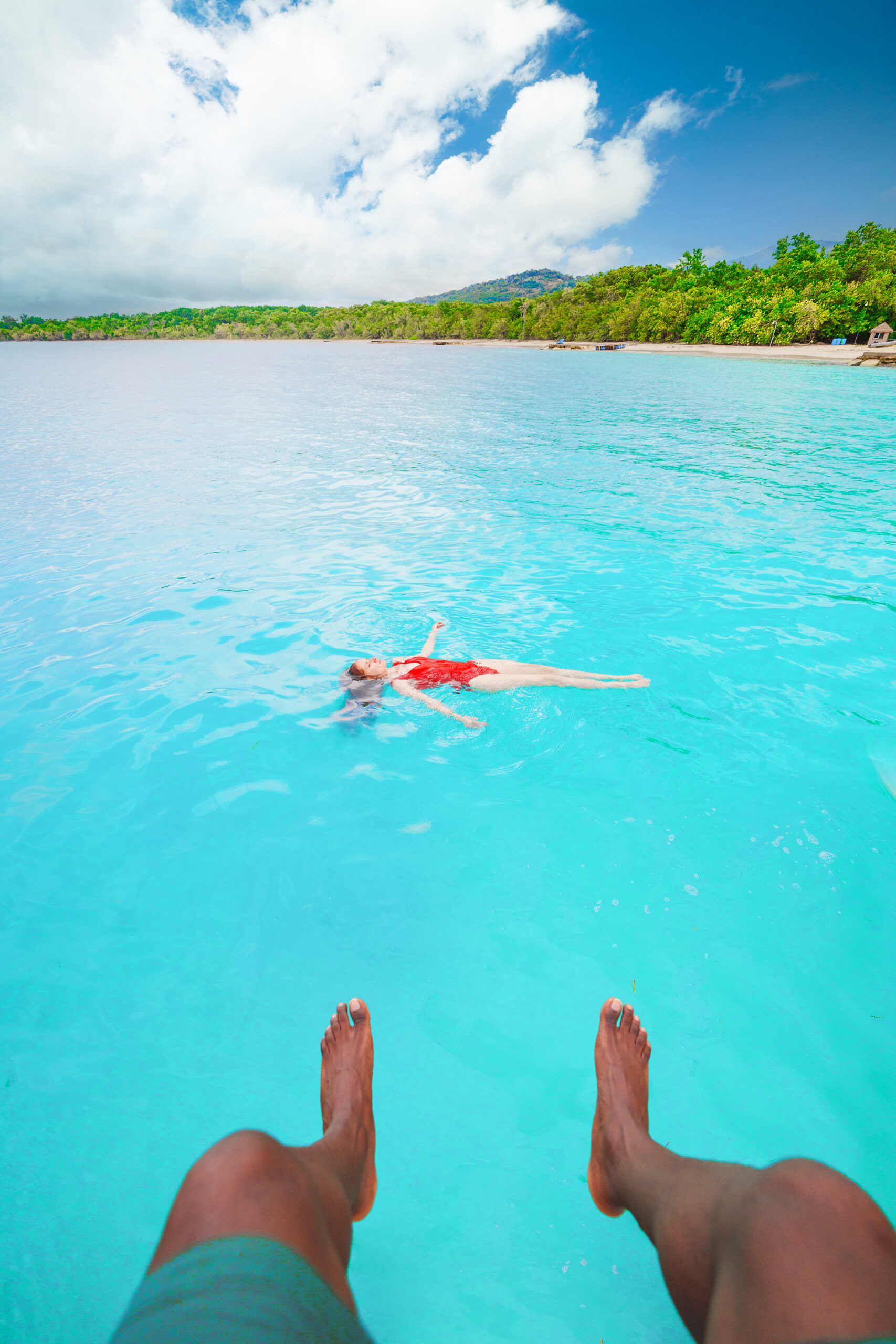 A man's legs dangling over the water with a woman floating on her back nearby